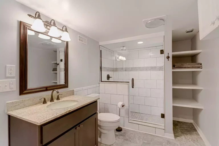 A white tiled bathroom with a stand-up shower, wood tone vanity and floating corner shelves.